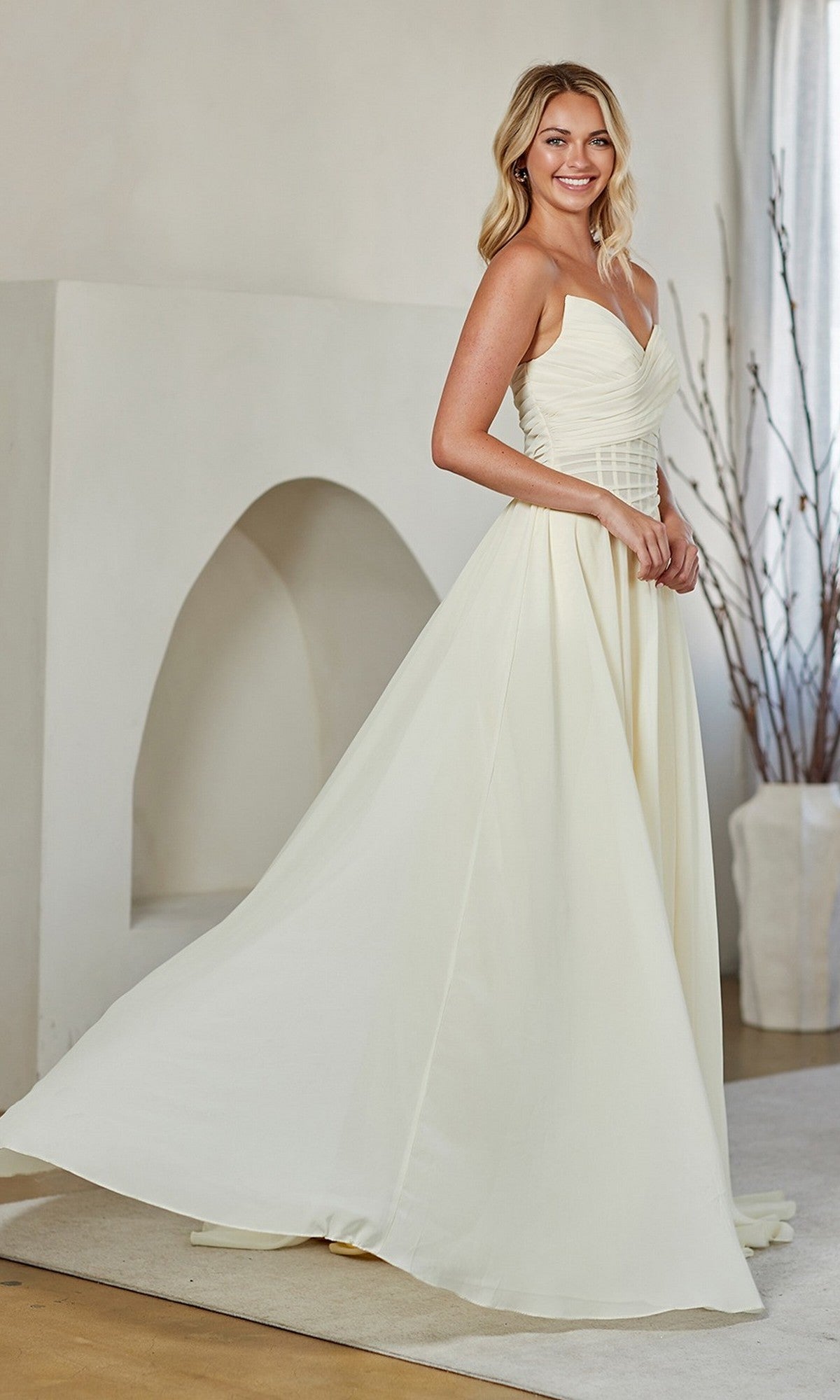A woman smiles indoors in the Amelia Couture 9097, a sleeveless, floor-length ivory prom dress with an elegant A-line skirt, holding one side to show its flowing style.